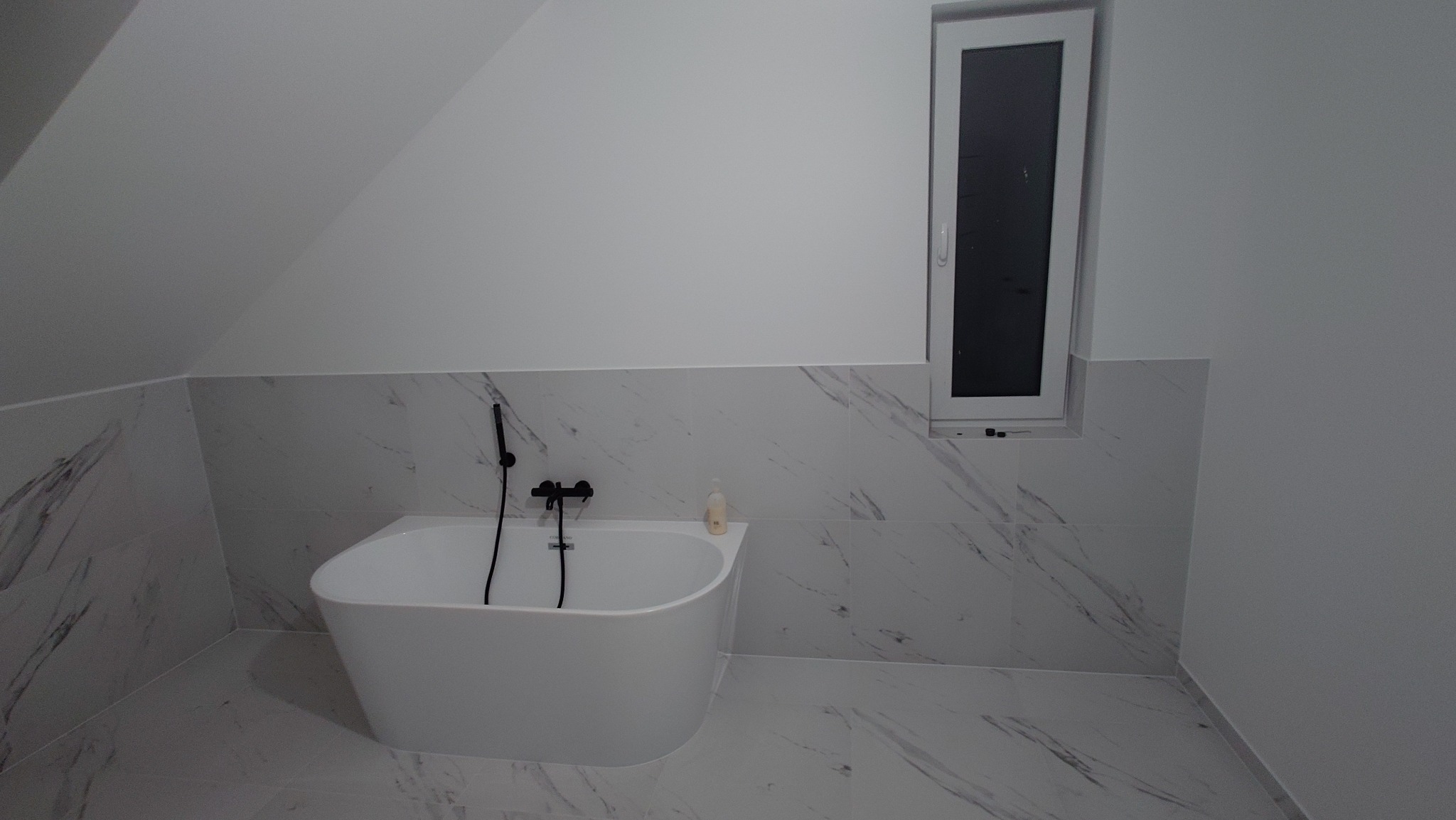 Biała, wolnostojąca wanna z czarną armaturą w łazience na poddaszu, wykończonej płytkami imitującymi marmur. W tle wąskie okno.