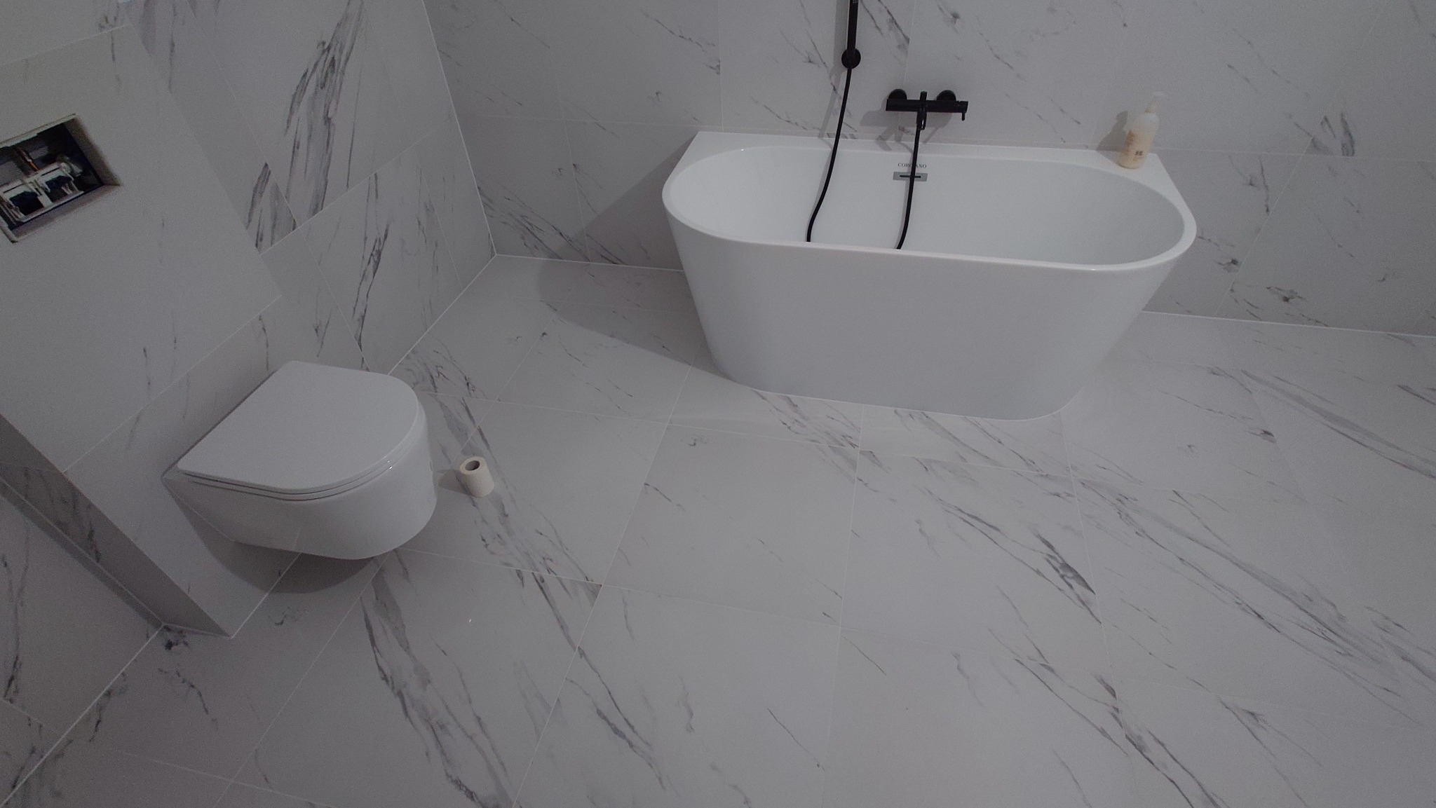 Nowoczesna łazienka z białą wanną wolnostojącą, podwieszaną toaletą i marmurowymi płytkami. Czarna armatura dodaje elegancji. Widok z góry.