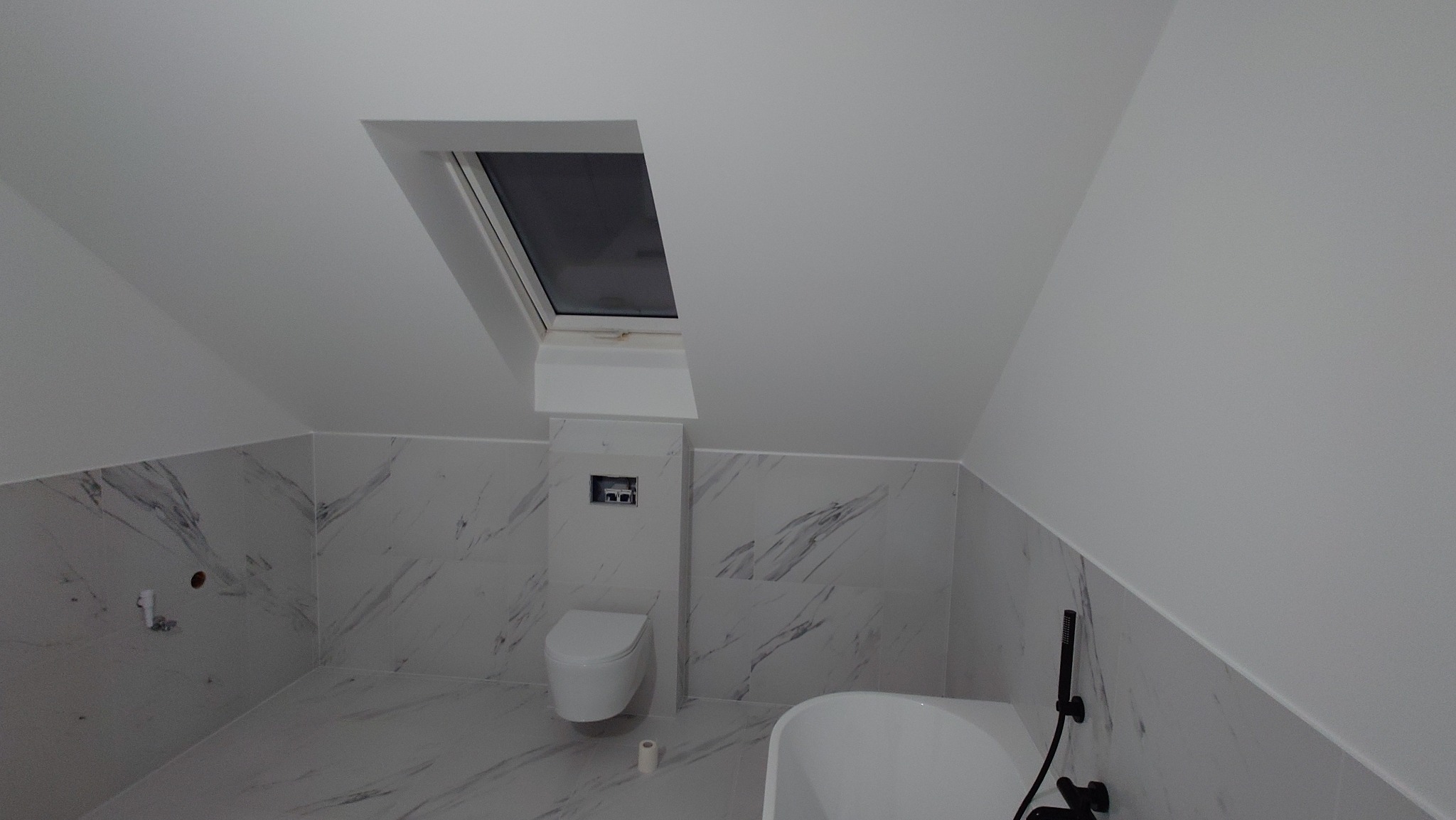 Wykończona łazienka na poddaszu z oknem dachowym. Białe płytki imitujące marmur na ścianach i podłodze. Wisząca toaleta i wanna. Minimalistyczny styl.