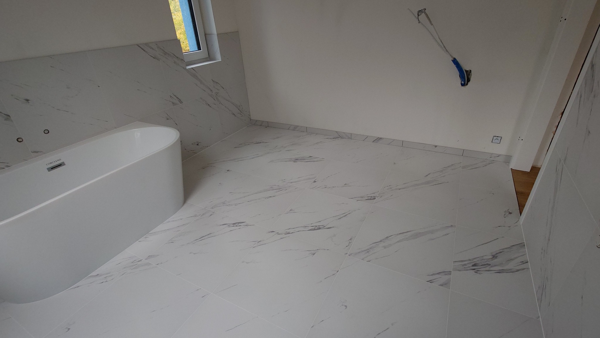 Biała, owalna wanna w rogu łazienki z marmurowymi płytkami na podłodze i ścianach. Widoczne gniazdko elektryczne i wystające kable. Okno w tle.