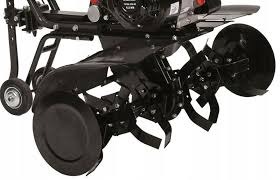 Część robocza glebogryzarki z ostrzami, widoczna z bliska, z elementami silnika u góry kadru. Oświetlenie studyjne, neutralne tło.