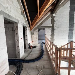 Smart Instal Szymon Juszczak - Wnętrze w trakcie instalacji elektrycznej: kable w peszlach ułożone wzdłuż ściany z białych bloczków, widoczne drewniane elementy konstrukcji dachu i fragment drewnianej balustrady, skrzynka...