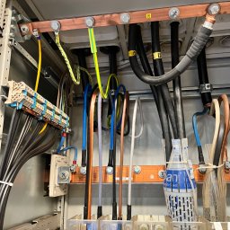 Smart Instal Szymon Juszczak - Wnętrze szafy elektrycznej z widocznymi przewodami w różnych kolorach, miedzianymi szynami zbiorczymi, bezpiecznikami i elementami łączeniowymi. Widoczne oznaczenia przewodów PE i L3.