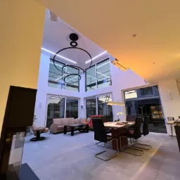 Nowoczesny salon z wysokim sufitem, oświetlony wiszącą lampą w kształcie okręgów, duże okna z widokiem na zewnątrz, minimalistyczny wystrój z beżową sofą i stołem jadalnym z krzesłami.