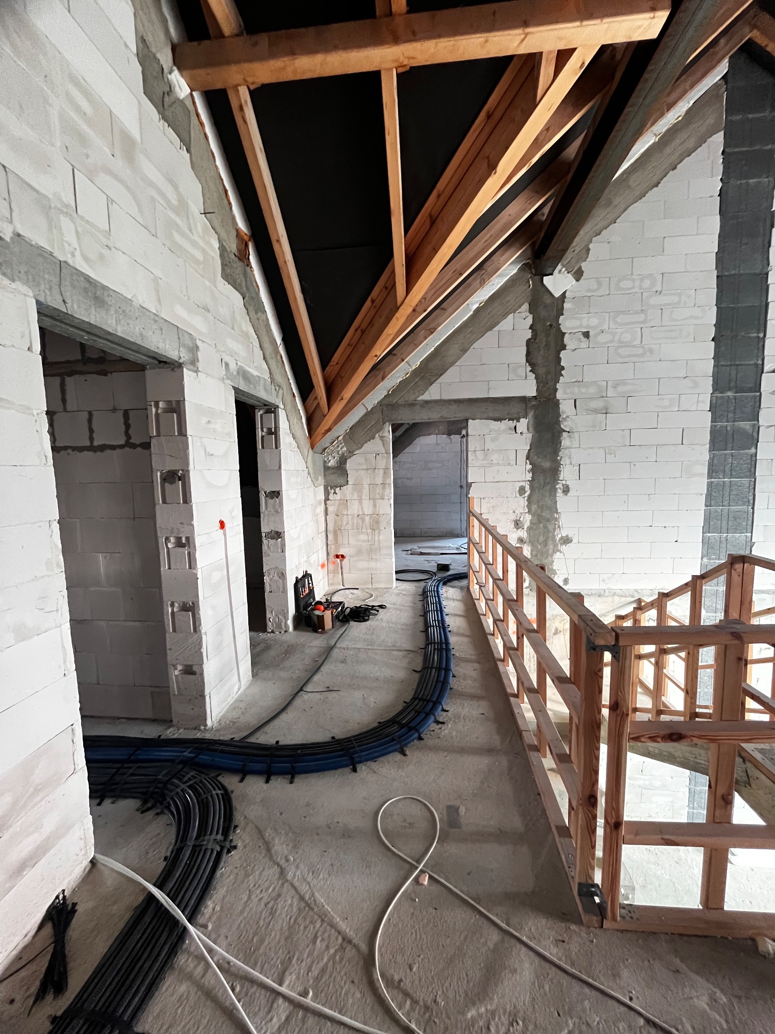 Wnętrze w trakcie instalacji elektrycznej: kable w peszlach ułożone wzdłuż ściany z białych bloczków, widoczne drewniane elementy konstrukcji dachu i fragment drewnianej balustrady, skrzynka...
