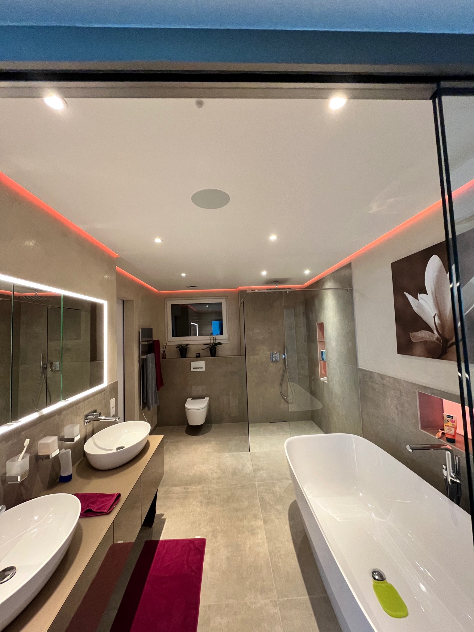 Nowoczesna łazienka z podświetleniem LED, dwiema umywalkami nablatowymi, wanną wolnostojącą i kabiną prysznicową typu walk-in, wykończona w szarościach i beżach, z akcentami bordowymi w postaci...