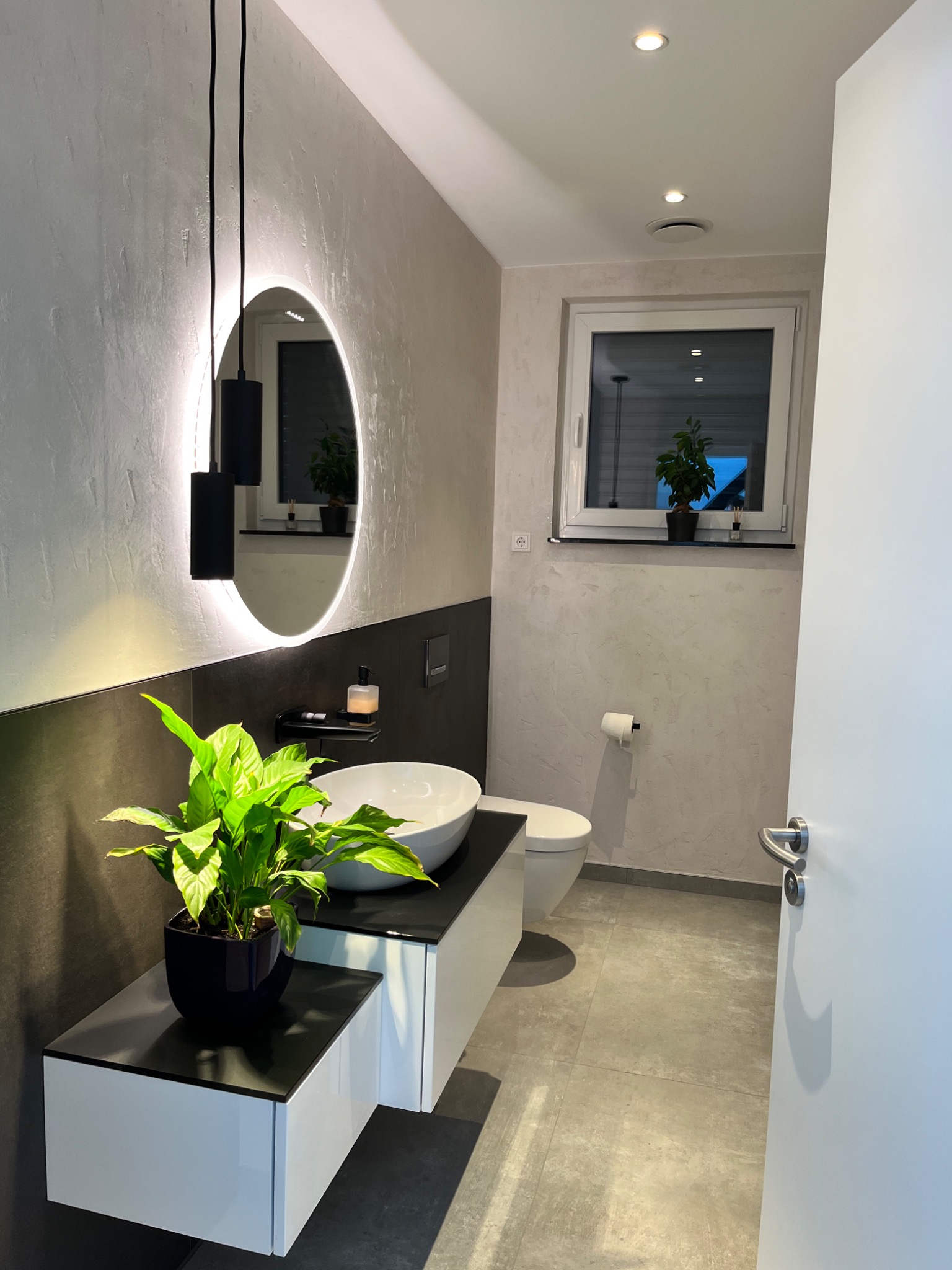 Nowoczesna łazienka z owalnym lustrem podświetlanym LED, wiszącą szafką z umywalką nablatową i rośliną doniczkową, wykończona szarą, teksturowaną ścianą i płytkami podłogowymi w odcieniach szarości.
