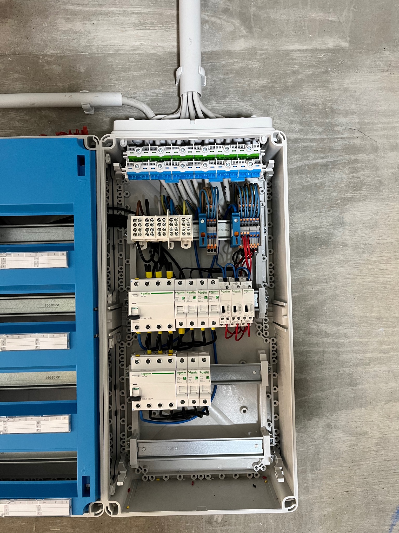 Otwarta skrzynka rozdzielcza z zainstalowanymi bezpiecznikami, przewodami i listwami zaciskowymi marki Schneider, umieszczona na szarej, betonowej ścianie.