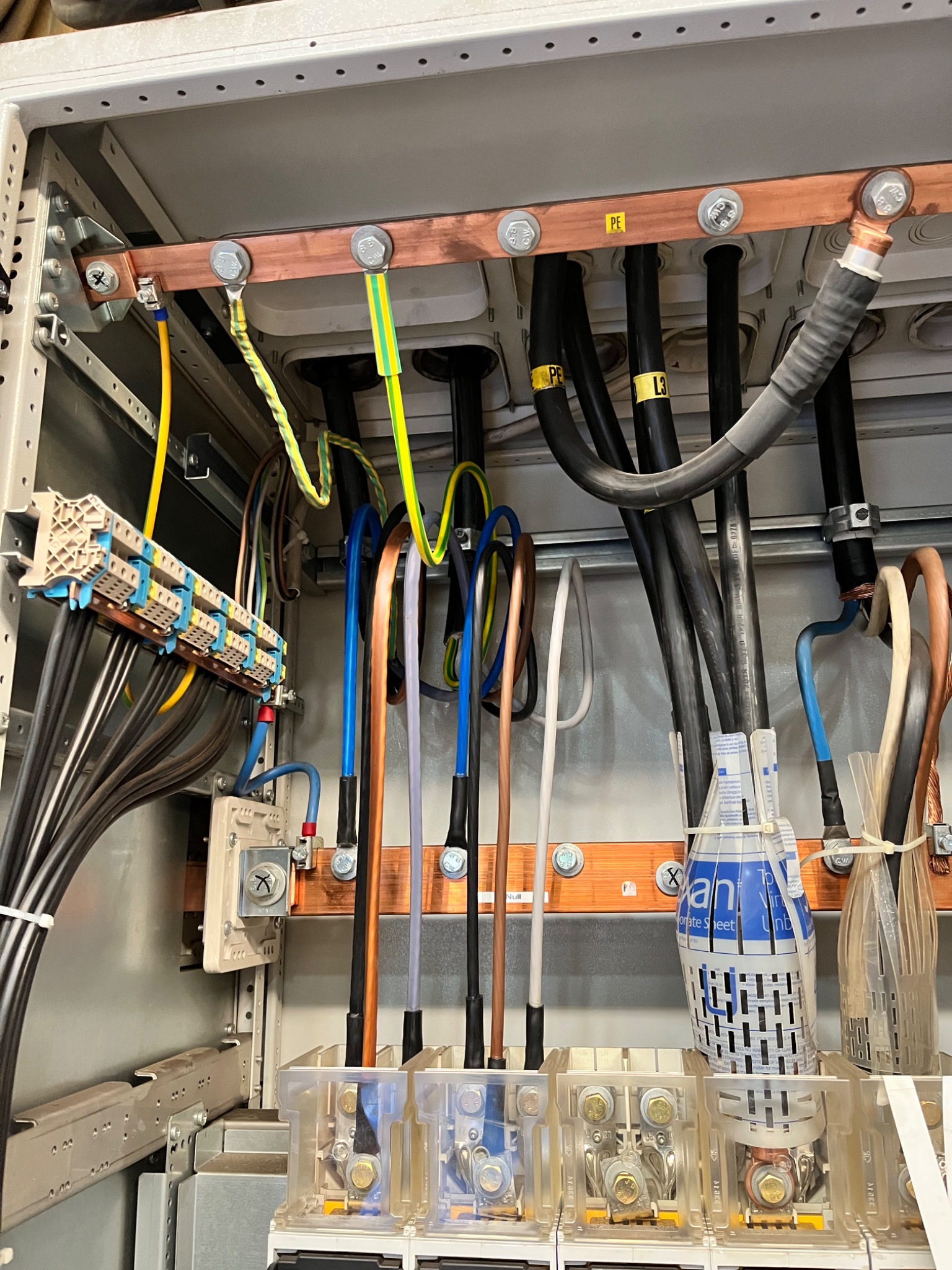 Wnętrze szafy elektrycznej z widocznymi przewodami w różnych kolorach, miedzianymi szynami zbiorczymi, bezpiecznikami i elementami łączeniowymi. Widoczne oznaczenia przewodów PE i L3.