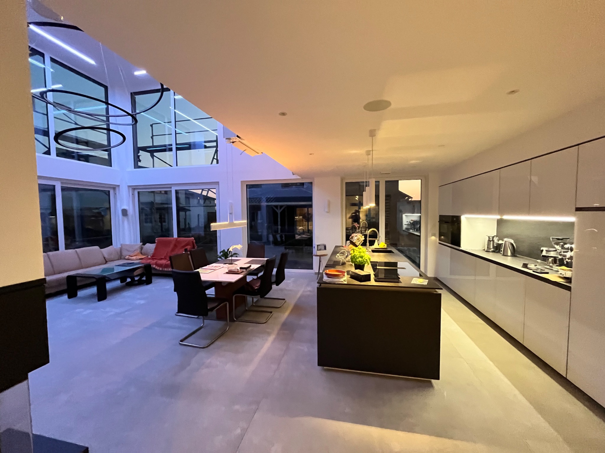 Przestronne, nowoczesne wnętrze domu z otwartą kuchnią i salonem, oświetlone ledowymi lampami sufitowymi i z dużymi oknami z widokiem na zewnątrz.