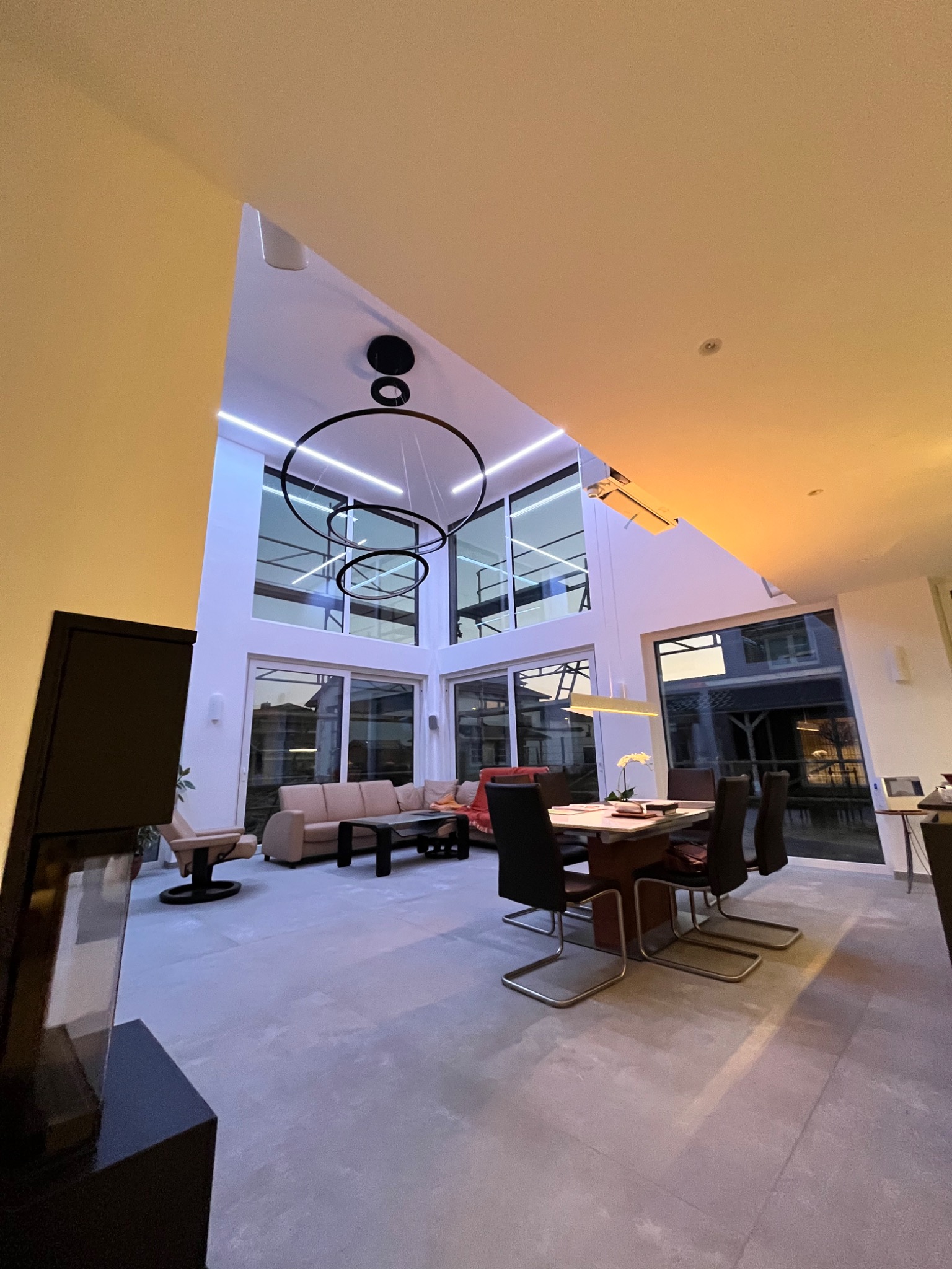 Nowoczesny salon z wysokim sufitem, oświetlony wiszącą lampą w kształcie okręgów, duże okna z widokiem na zewnątrz, minimalistyczny wystrój z beżową sofą i stołem jadalnym z krzesłami.