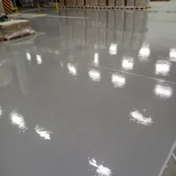 Gładka, szara powierzchnia w hali magazynowej w Sosnowcu, odbijająca światło lamp sufitowych i regały z zapakowanymi towarami.