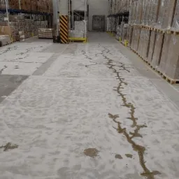 Naprawa pęknięć w posadzce betonowej magazynu, wypełnione szarą masą, widoczne palety z towarem w tle.