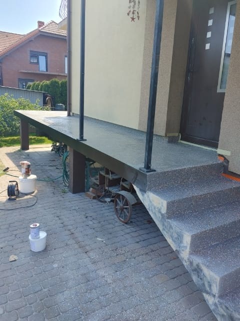 Zewnętrzny taras i schody pokryte szarą, ziarnistą nawierzchnią, widoczne puszki z farbą i wózek na cegły w tle.