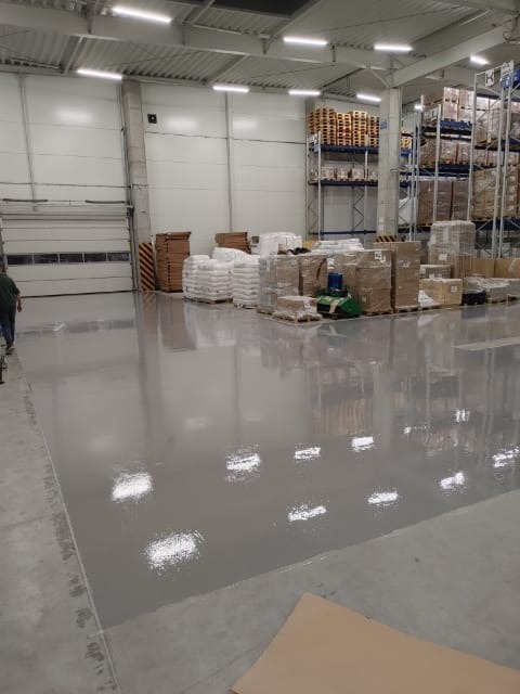 Błyszcząca, szara podłoga w magazynie przemysłowym, odbijająca światła sufitowe, z paletami i towarami w tle.