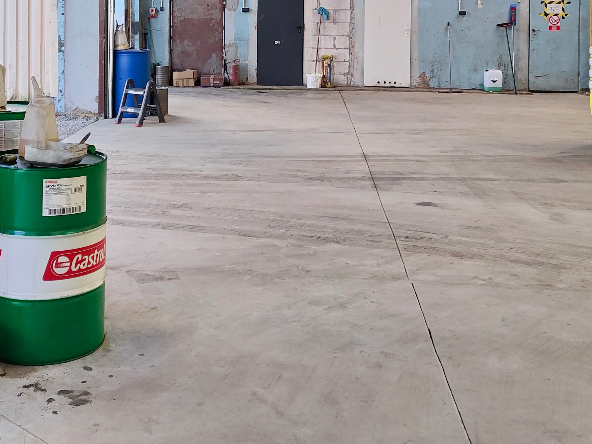 Szara, betonowa posadzka w hali przemysłowej z widocznymi śladami użytkowania, zielona beczka Castrol z resztkami żywicy i narzędziami w tle, różne drzwi i akcesoria na ścianie.