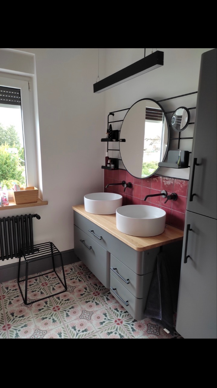 Nowoczesna łazienka z dwoma okrągłymi umywalkami nablatowymi, szarą szafką z drewnianym blatem, okrągłym lustrem na tle czerwonej mozaiki i wzorzystą podłogą w stylu retro.