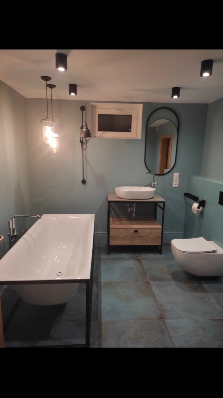 Nowoczesna łazienka z wolnostojącą wanną na metalowym stelażu, umywalką nablatową na szafce z drewnianymi elementami, wiszącą toaletą i niebiesko-szarymi płytkami podłogowymi, oświetlona lampami...