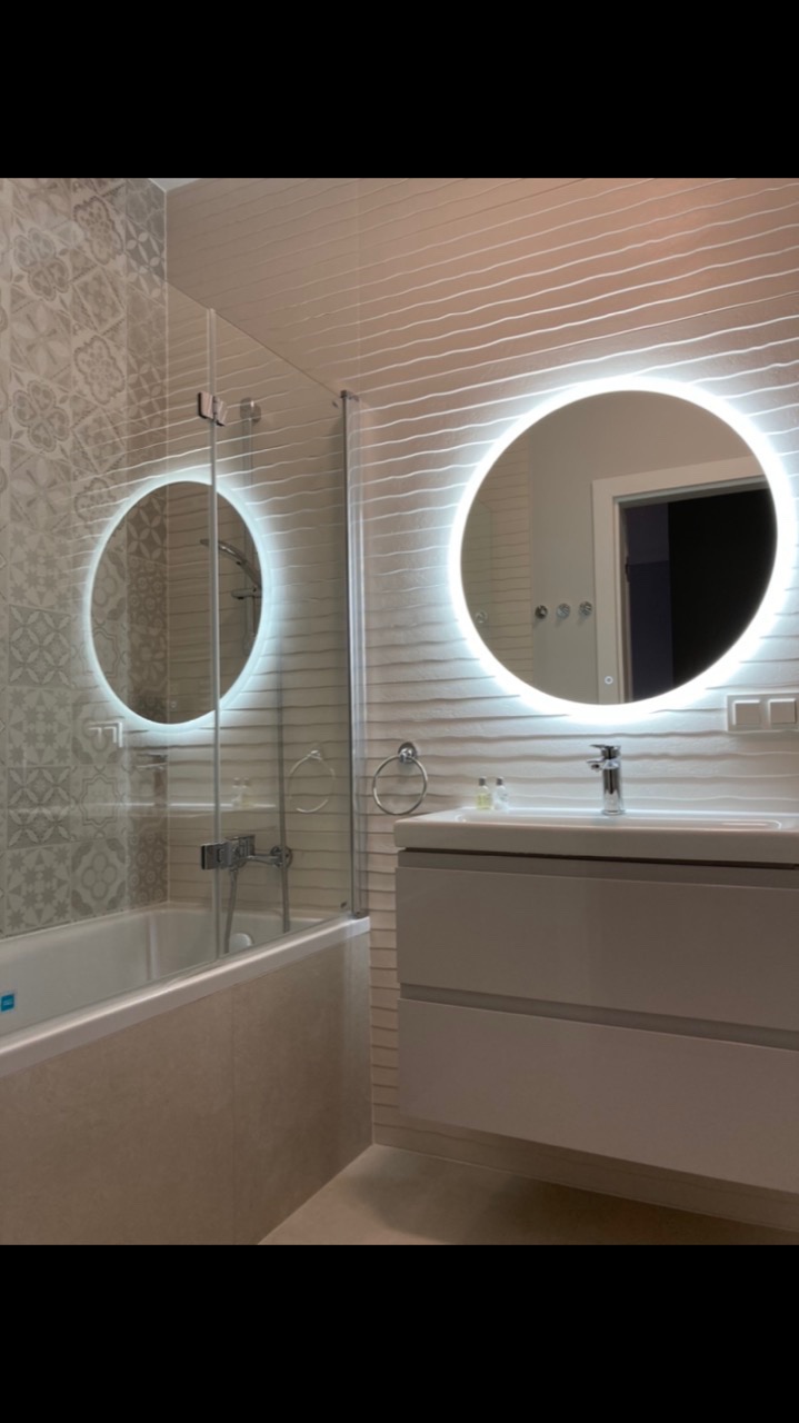 Nowoczesna łazienka z wanną za szklaną ścianką, okrągłym lustrem z podświetleniem LED, umywalką z szafką w kolorze złamanej bieli i dekoracyjnymi płytkami na ścianach.