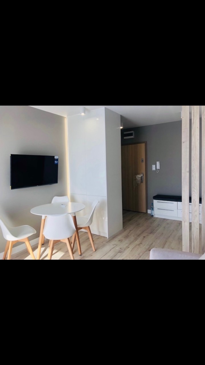 Jasne wnętrze mieszkania po remoncie: biały okrągły stół z trzema krzesłami, telewizor na ścianie, drewniana podłoga i elementy dekoracyjne z drewna.