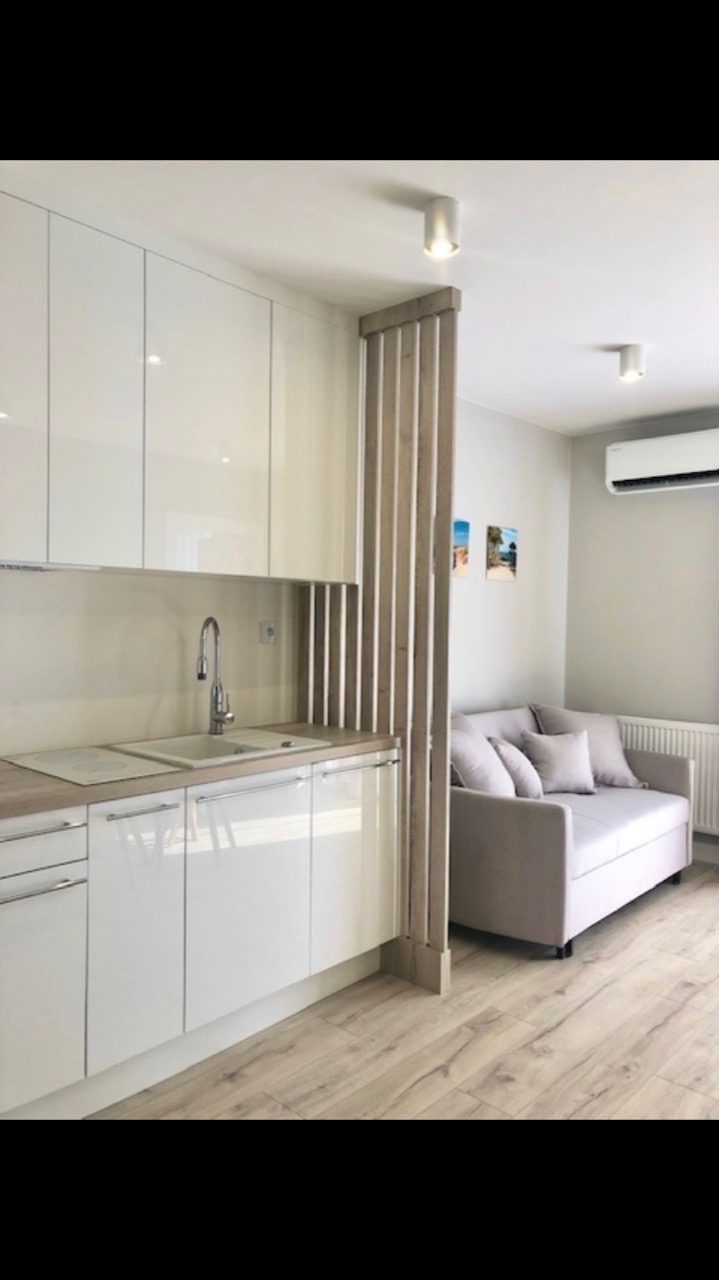 Nowoczesna, jasna kuchnia z białymi, lakierowanymi szafkami i drewnianą przegrodą, za którą widać szarą sofę w minimalistycznym stylu.