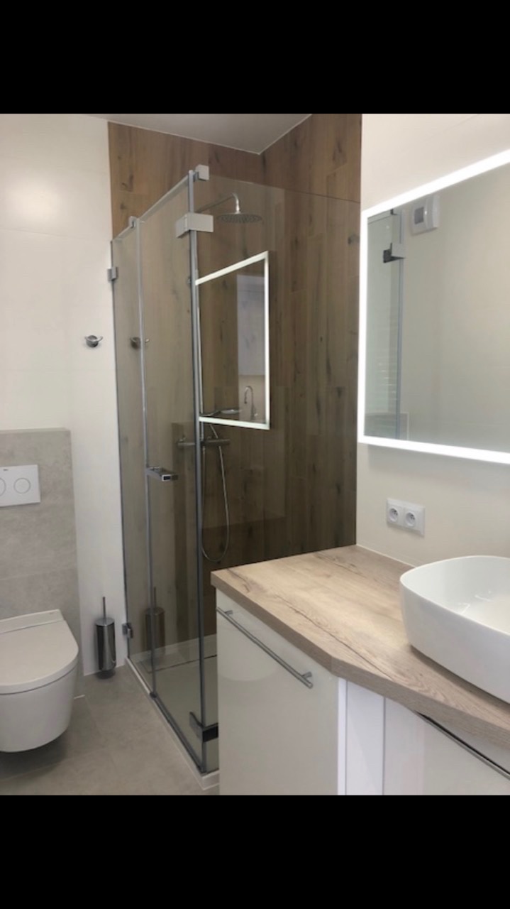 Nowoczesna łazienka z kabiną prysznicową typu walk-in, umywalką nablatową i lustrem z podświetleniem LED, wykończona płytkami imitującymi drewno i szarą ceramiką.