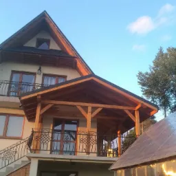 Dwupiętrowy dom z drewnianą werandą i kutą balustradą, widok z dołu na tle błękitnego nieba, obok szklarnia.