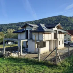 Budowa domu z betonu komórkowego w stanie surowym otwartym, z widoczną konstrukcją dachu pokrytą ciemną dachówką, na tle zielonego wzgórza i błękitnego nieba.