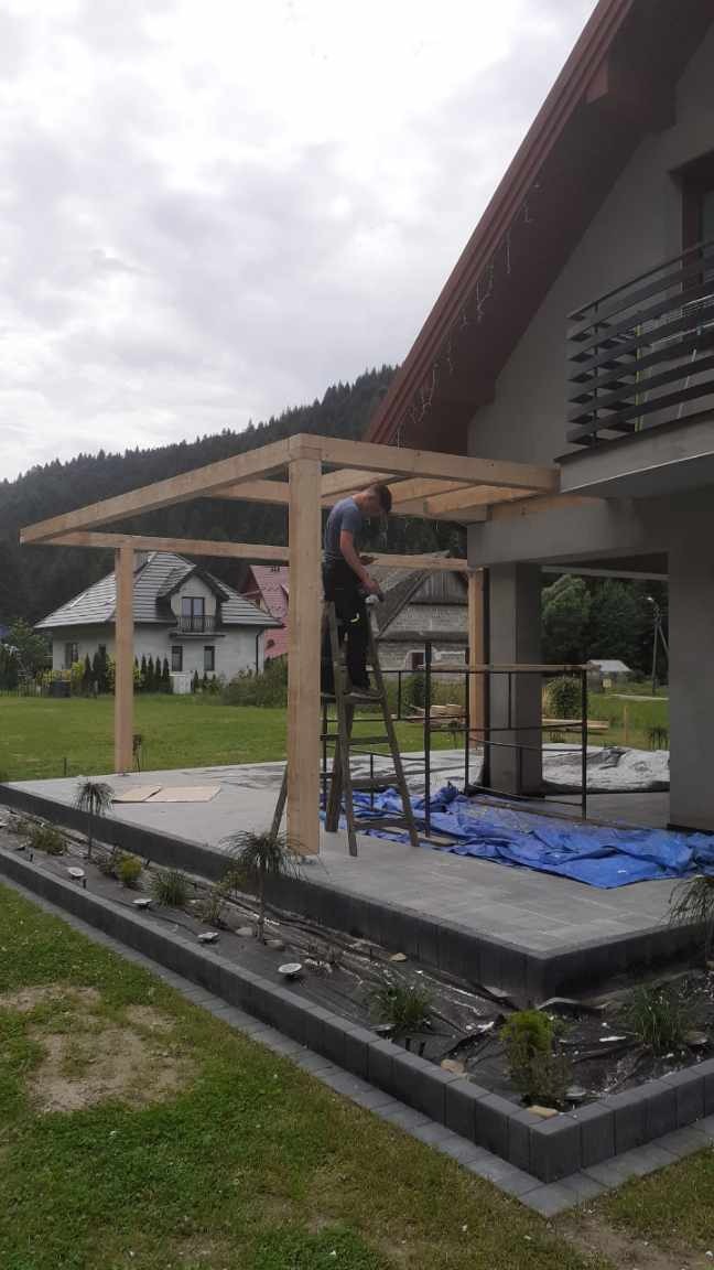 Montaż drewnianej konstrukcji z belek na tarasie domu jednorodzinnego, widoczny pracownik na drabinie podczas pracy, w tle budynki mieszkalne i zalesione wzgórza.