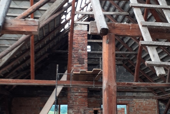 Konstrukcja dachu w trakcie remontu z widoczną więźbą dachową, kominem z czerwonej cegły i fragmentami ściany, rusztowanie wewnątrz budynku.