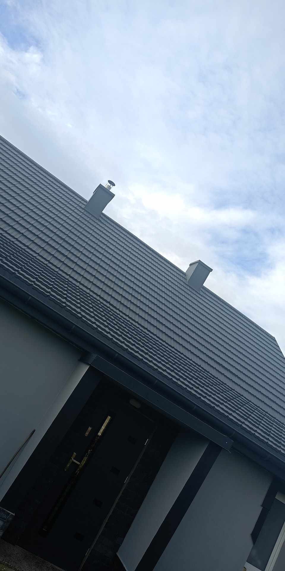 Nowoczesny dom z ciemnoszarą dachówką ceramiczną i dwoma kominami, widok z dołu na tle pochmurnego nieba.