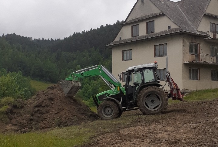 Zielony traktor z łyżką nabiera ziemię na tle budynku i zalesionych wzgórz. Prace ziemne w toku, przygotowanie terenu pod budowę lub zagospodarowanie.