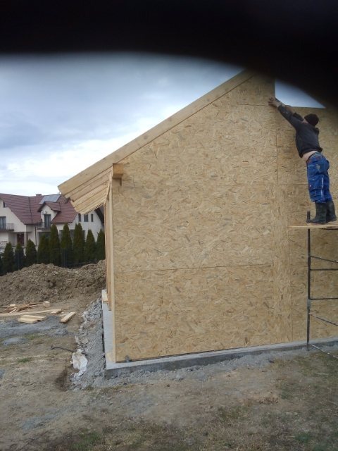 Budowa domu szkieletowego: montaż płyt OSB na konstrukcji, widoczny pracownik na rusztowaniu, częściowo ułożona więźba dachowa, w tle domy jednorodzinne.