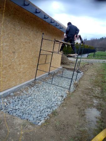 Pracownik na rusztowaniu przy ścianie domu szkieletowego z płyty OSB, widoczne orynnowanie, podsypka z kamienia i częściowo wykonana elewacja.