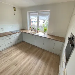 Jasna kuchnia na wymiar z drewnianym blatem, fronty szafek w odcieniach bieli, złote uchwyty, okno z roletami rzymskimi, drewniana podłoga, widok z perspektywy narożnika.