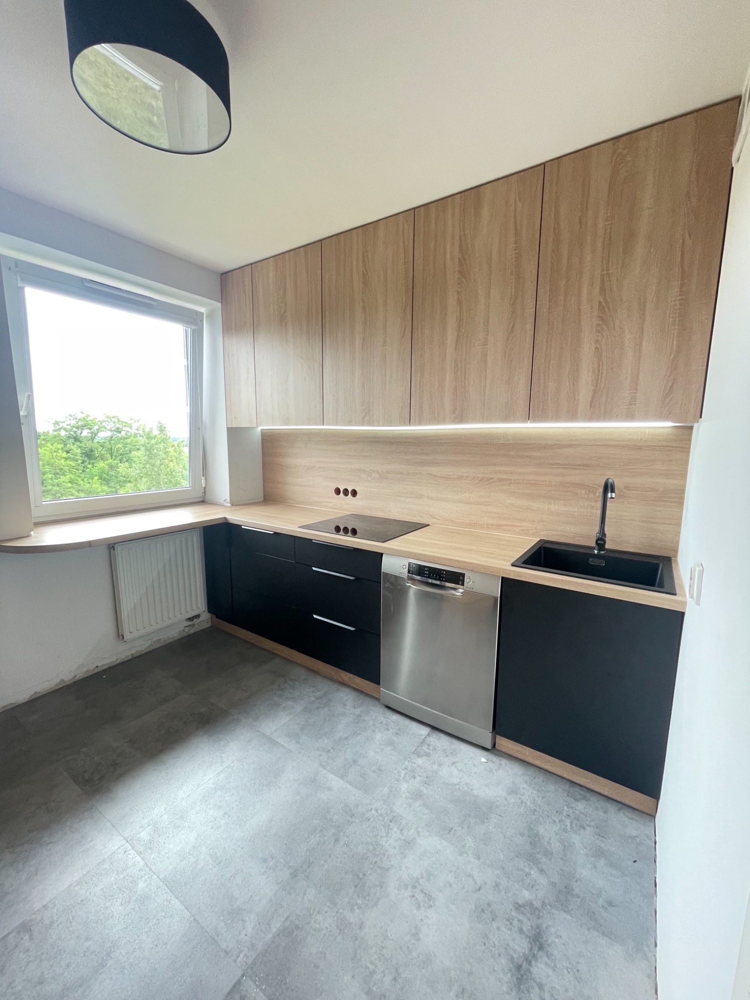 Nowoczesna kuchnia z drewnianymi szafkami górnymi, czarnymi szafkami dolnymi, blatem w kolorze drewna i szarą podłogą; widoczny fragment okna z widokiem na zieleń.