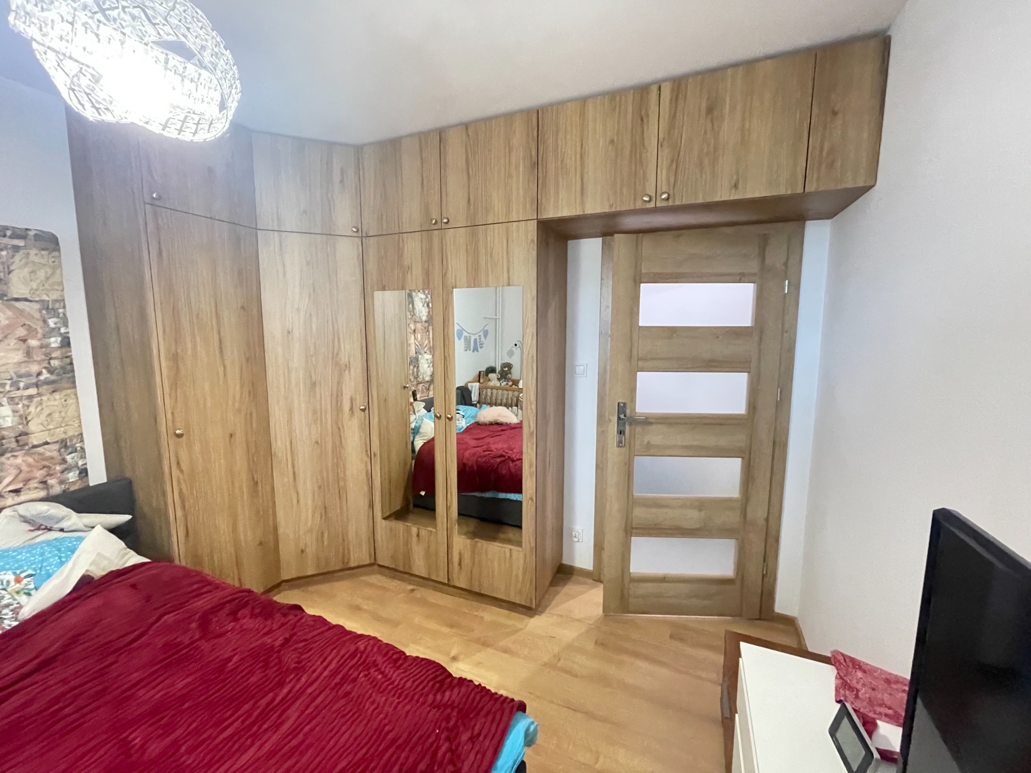 Drewniana szafa narożna z lustrami w Toruniu, zabudowa od podłogi do sufitu, widoczna lampa sufitowa i fragment sypialni z czerwonym nakryciem łóżka.