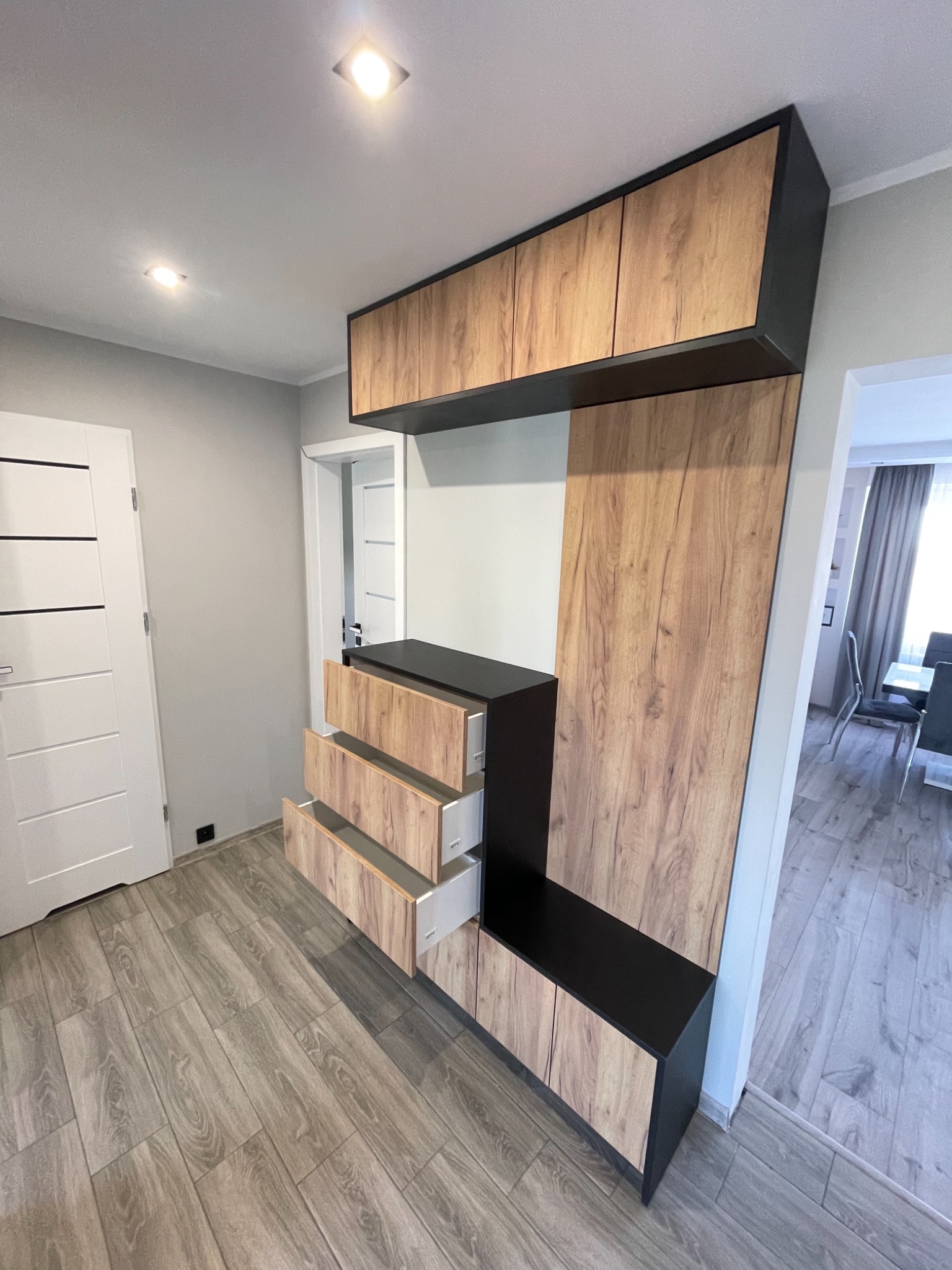 Zabudowa meblowa z otwartymi szufladami i szafkami wiszącymi, fronty w odcieniach drewna, korpus w kolorze czarnym, widok na wnętrze mieszkania z podłogą imitującą drewno.
