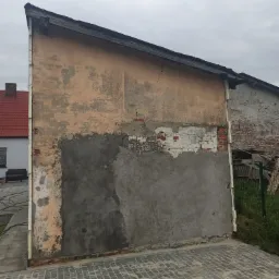 Fragment ściany budynku z widocznymi śladami uszkodzeń i napraw, częściowo pokryty tynkiem, z odsłoniętymi fragmentami cegieł i starej farby, na tle kostki brukowej i sąsiednich budynków.