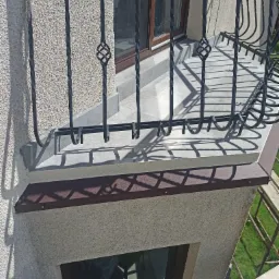 Wykończony balkon z kutą, czarną balustradą, marmurowymi płytkami na posadzce i brązowym wykończeniem krawędzi, widok z góry
