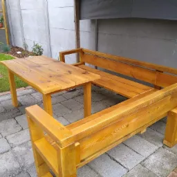 Drewniany stół z ławkami w ogrodzie na brukowanym patio, z widocznymi elementami roślinności i betonowym ogrodzeniem w tle.