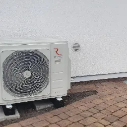 Biała zewnętrzna jednostka pompy ciepła Rotenso Hiro Series zamontowana na betonowych bloczkach przy ścianie domu z kostki brukowej.