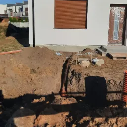 Wykop pod rury kanalizacyjne na działce budowlanej, widoczne zabezpieczenia taśmą ostrzegawczą, w tle dom z brązowymi roletami i częściowo zakrytymi drzwiami wejściowymi.