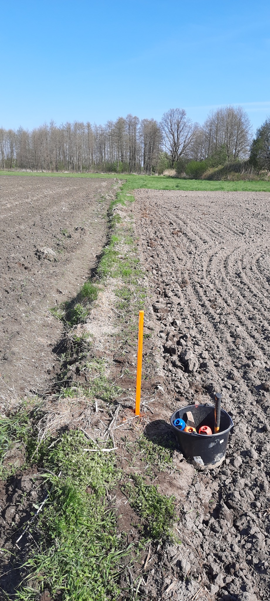 Pomarańczowy palik wbity w ziemię, wyznaczający granicę na zaoranym polu. Obok czarne wiadro z narzędziami i kolorowymi znacznikami.