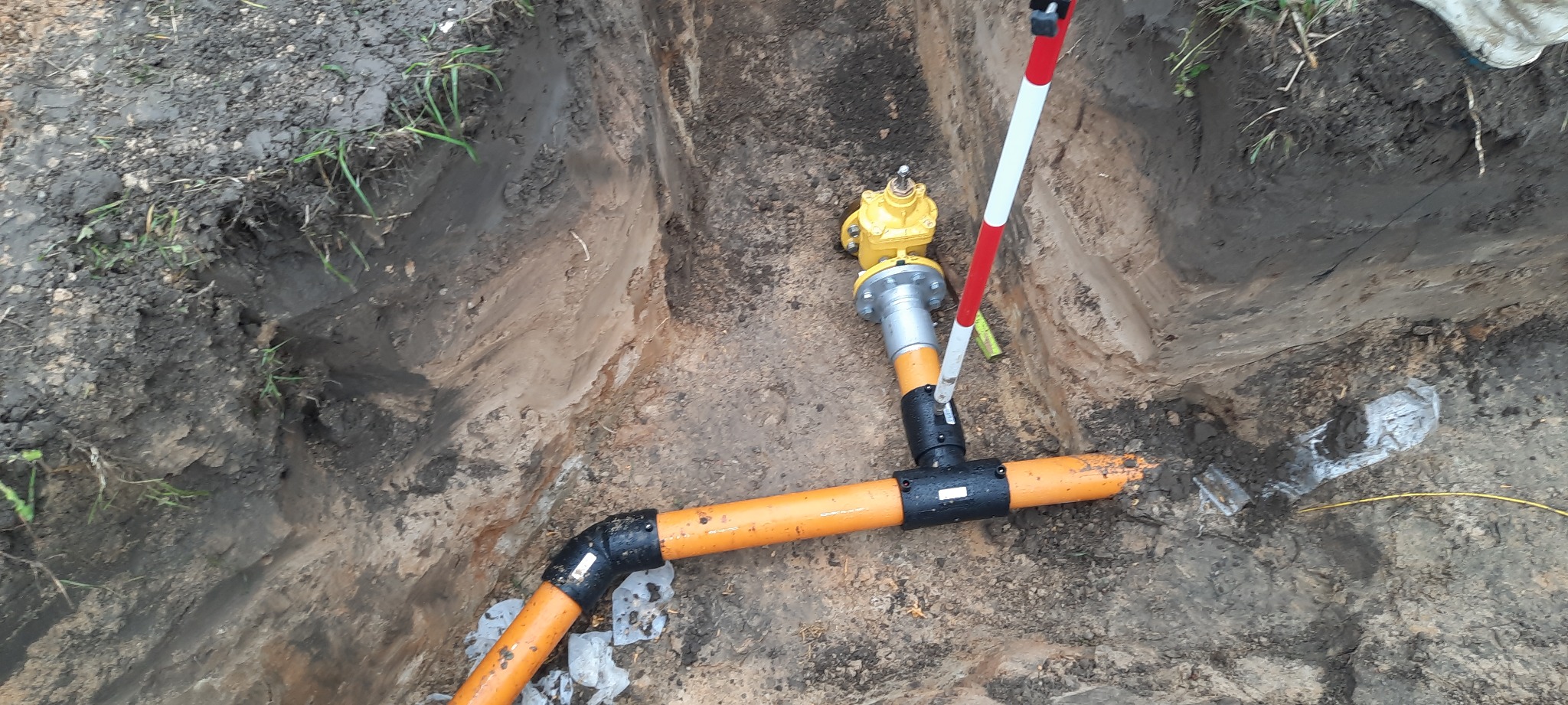 Wykop z pomarańczowymi rurami wodociągowymi, żółty zawór i tyczka geodezyjna z białymi i czerwonymi paskami.
