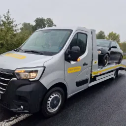 Srebrny samochód pomocy drogowej Renault z czarnym autem osobowym na lawecie, stojący na mokrej nawierzchni drogi w pochmurny dzień.