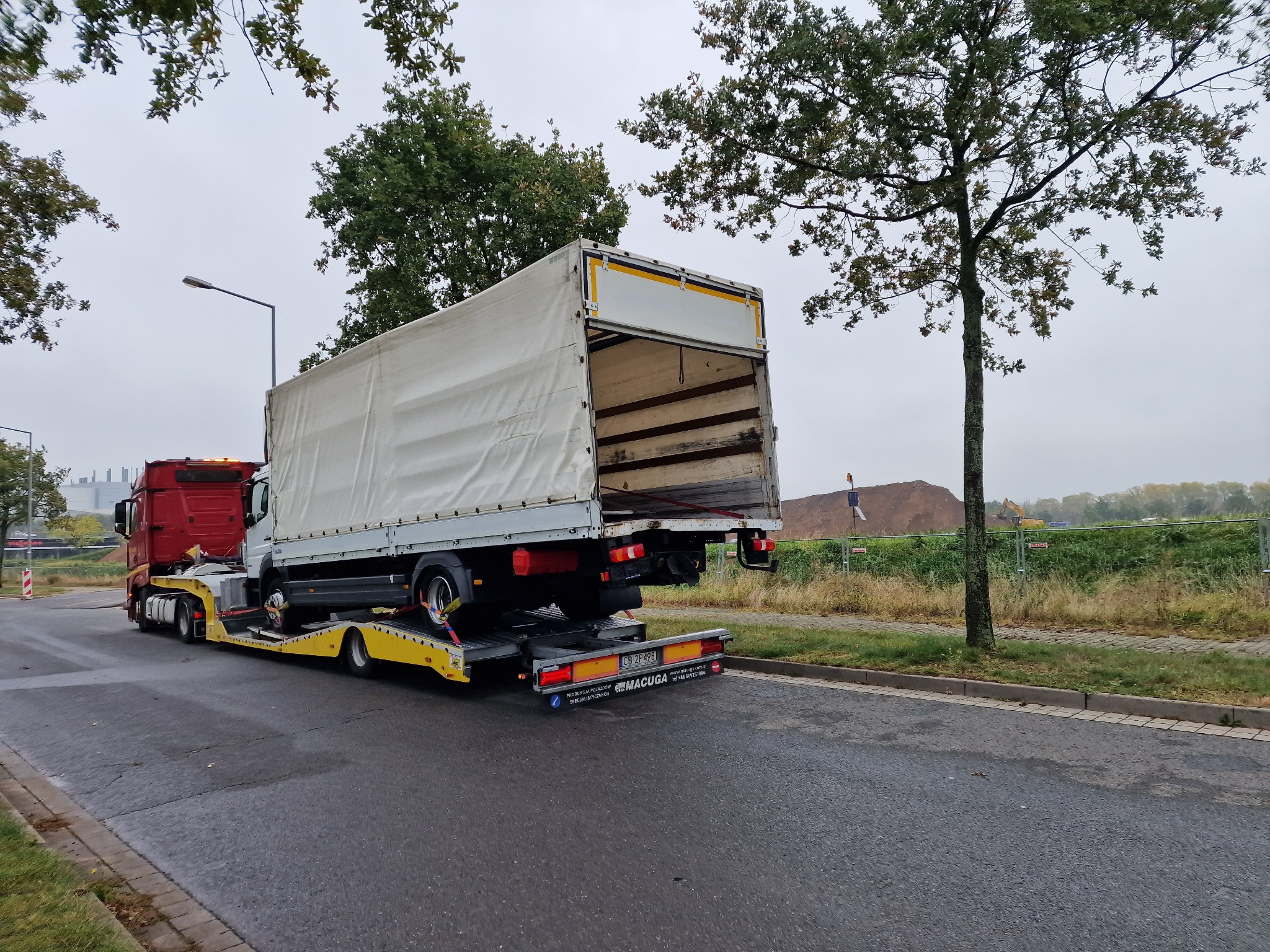 Czerwona laweta z żółtymi elementami transportuje białą ciężarówkę z otwartą plandeką na tle szarego nieba i drzew.