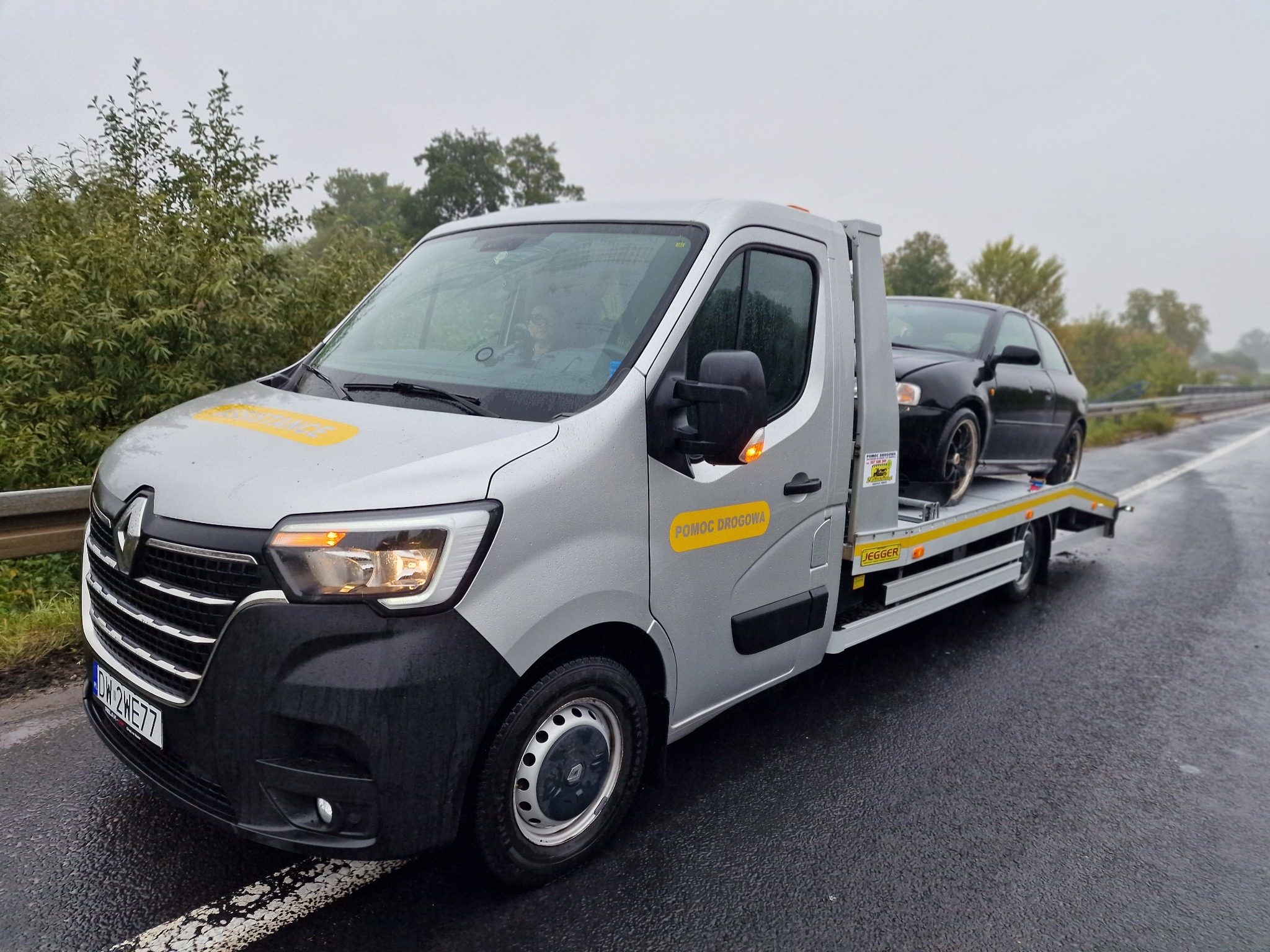 Srebrny samochód pomocy drogowej Renault z czarnym autem osobowym na lawecie, stojący na mokrej nawierzchni drogi w pochmurny dzień.