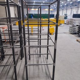 BAJ-DESIGN Błażej Dostatni - Metalowe szkielety konstrukcji spawane w hali produkcyjnej, widoczna osoba w kombinezonie spawalniczym w tle.