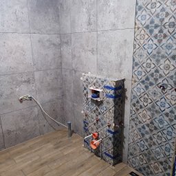 BAJ-DESIGN Błażej Dostatni - Wnętrze łazienki w trakcie remontu: położone płytki imitujące drewno na podłodze, częściowo wyłożone płytki na ścianach, w tym jedna ściana z wzorem kwiatowym, widoczne elementy instalacji...